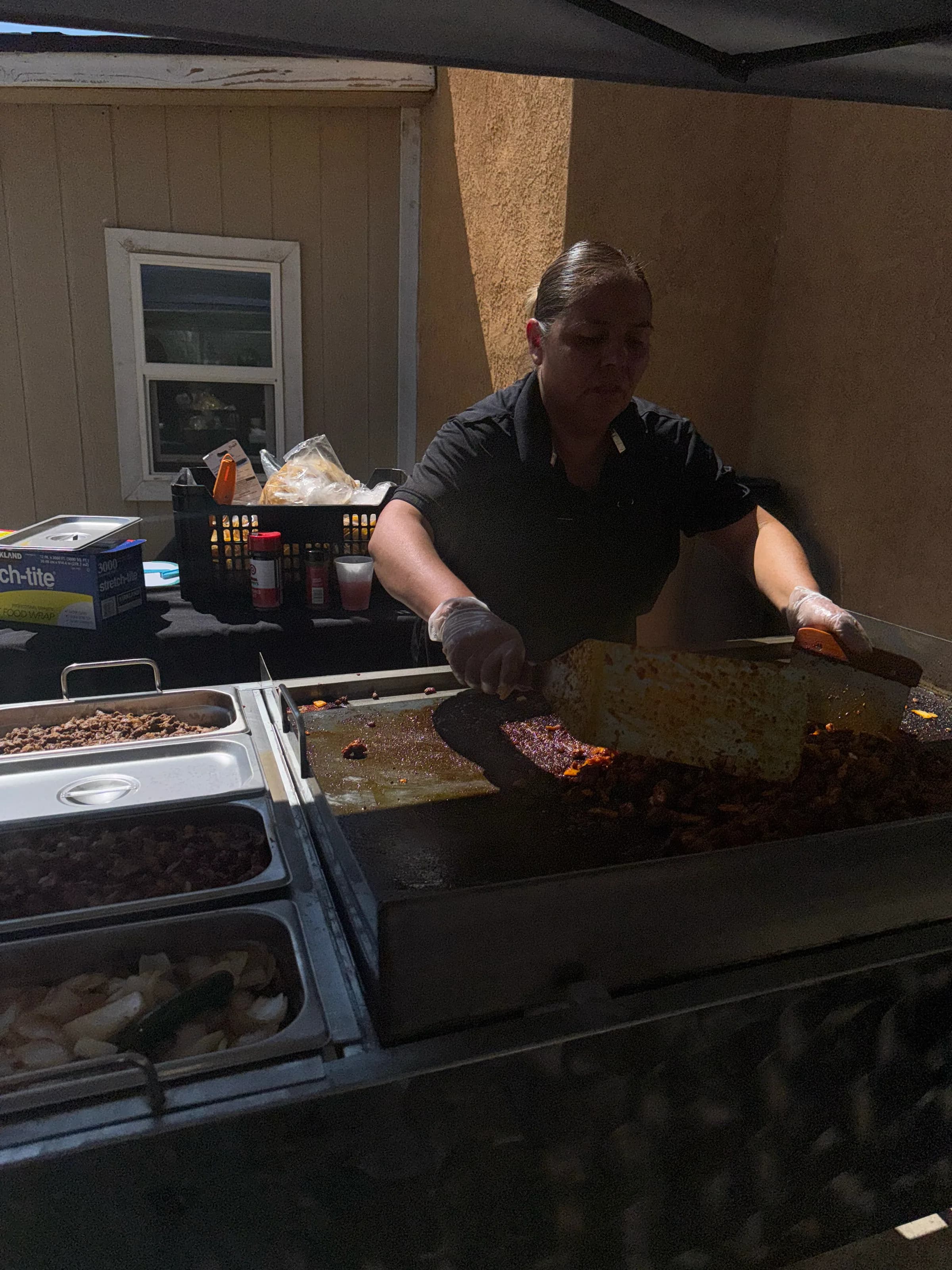 Tacos V's owner working the flat-top grill with two spatulas, cooking al pastor at an outdoor catering event