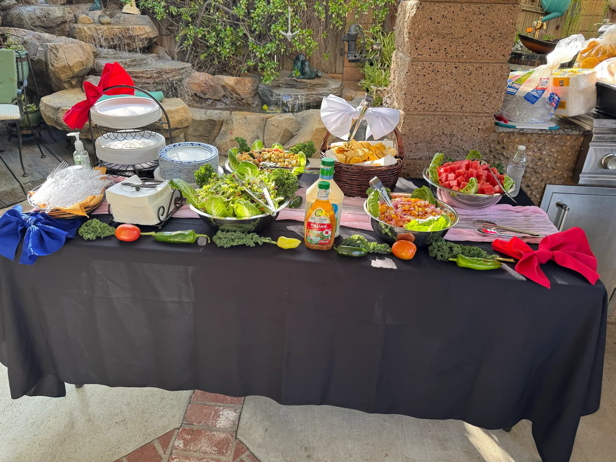 Elegant Tacos V sides table at a quinceañera, green salad, pasta salad, fresh fruit, and chips against a stone garden backdrop