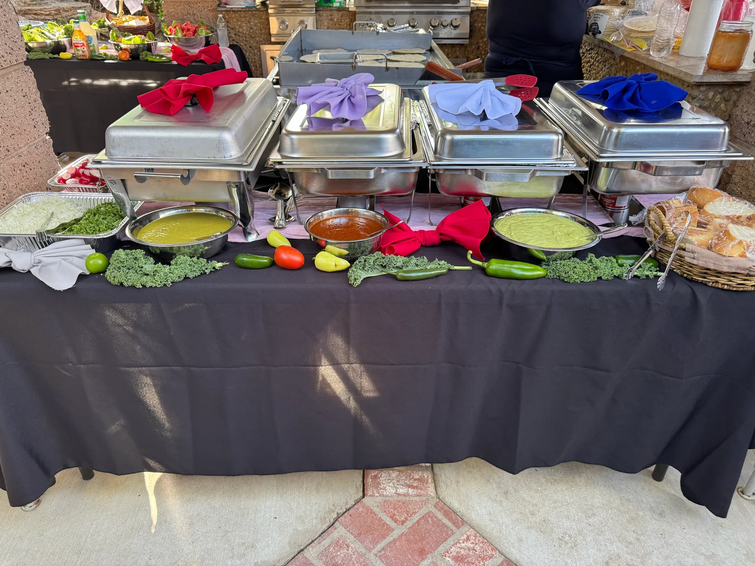 Tacos V sides station, four chafing dishes with colored bow accents, three house-made salsas, and bread, all set on a black tablecloth at a backyard catering event